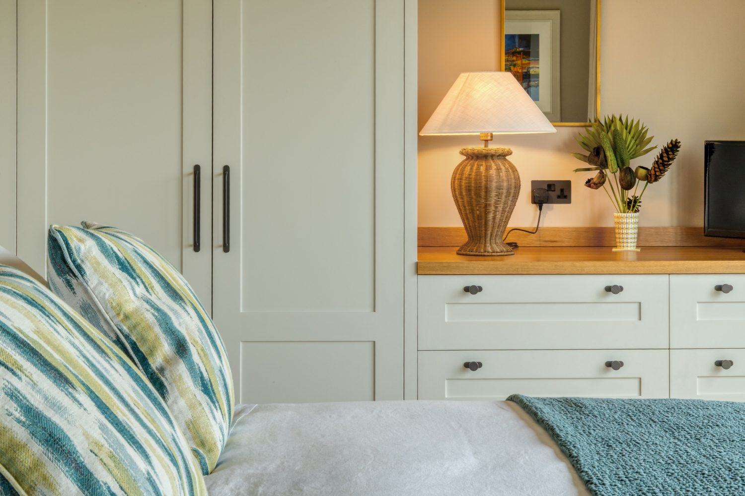 Edge of bed with green cushions with cream wardrobe and drawers behind with wicker lamp and plant on top