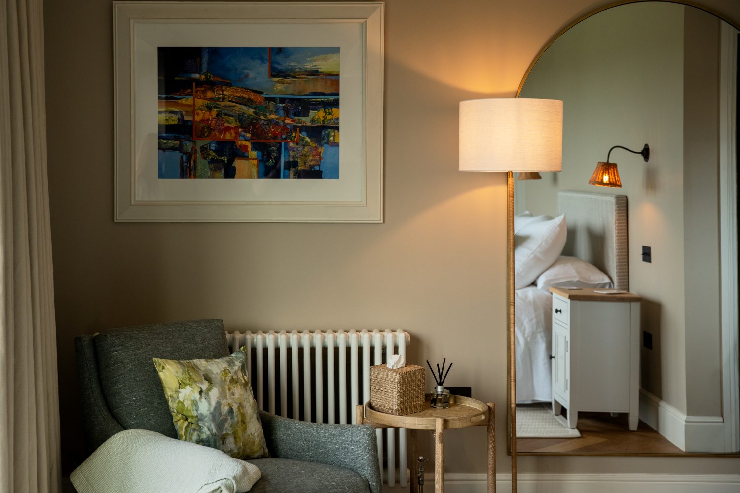 Corner of bedroom with large framed artwork on wall beside standing lamp and large arched mirror. Grey chair and wooden side table below