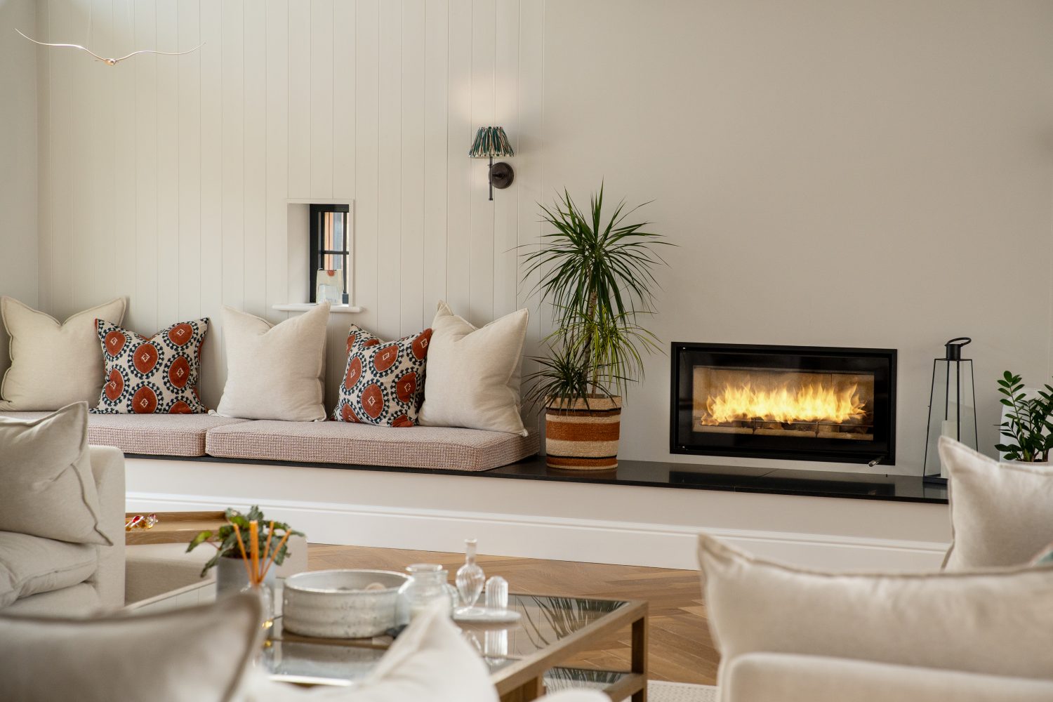 Cushioned bench seating area to the left of leafy plant and black fireplace built into cream panelled wall