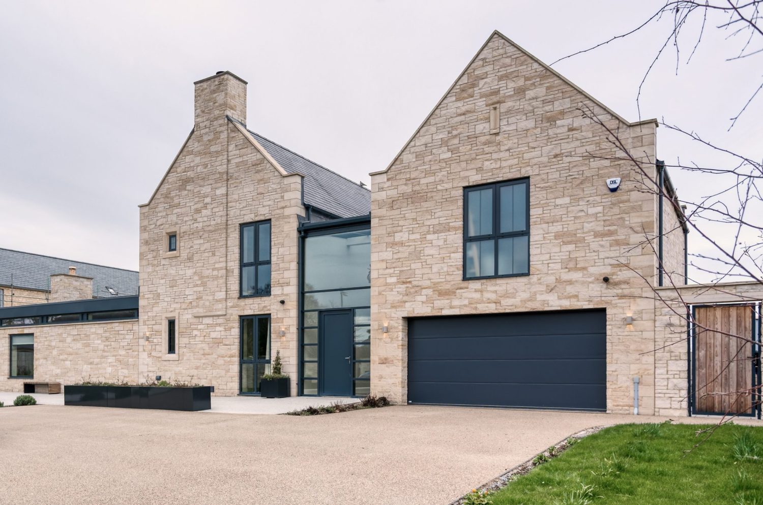 Exterior front of house with exposed stone walls, large black framed windows, black garage door and large driveway in front